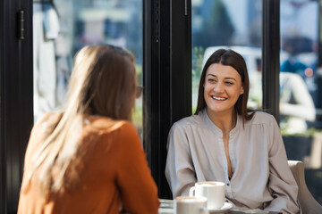 Two young business women in a cafe having one on one meeting. Friends after work talking gossiping and having coffee at a window table on a sunny day..