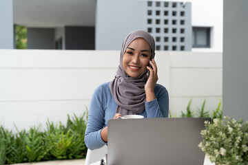 Wall Mural - Portrait of Muslim asian woman in hijab using laptop while taking call phone at home.