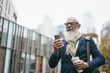Business hipster senior man using mobile phone while walking to work with city in background - Focus face