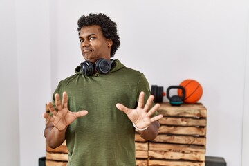 Poster - African man with curly hair wearing sportswear at the gym afraid and terrified with fear expression stop gesture with hands, shouting in shock. panic concept.