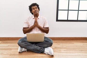 Wall Mural - African man with curly hair using laptop sitting on the floor begging and praying with hands together with hope expression on face very emotional and worried. begging.