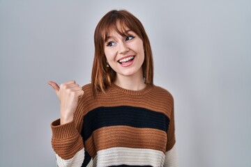 Poster - Young beautiful woman wearing striped sweater over isolated background smiling with happy face looking and pointing to the side with thumb up.