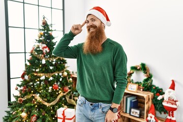 Sticker - Redhead man with long beard wearing christmas hat by christmas tree smiling pointing to head with one finger, great idea or thought, good memory