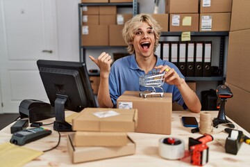 Sticker - Young man working at small business ecommerce holding small cart pointing thumb up to the side smiling happy with open mouth