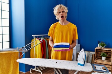 Poster - Young man ironing clothes at home in shock face, looking skeptical and sarcastic, surprised with open mouth