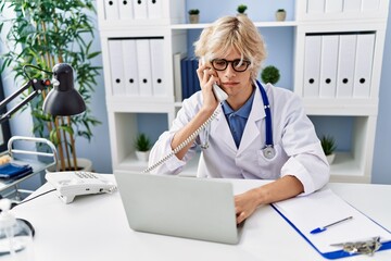 Poster - Young blond man doctor using laptop talking on telephone at clinic