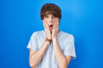 Canvas Print - Hispanic young man standing over blue background afraid and shocked, surprise and amazed expression with hands on face