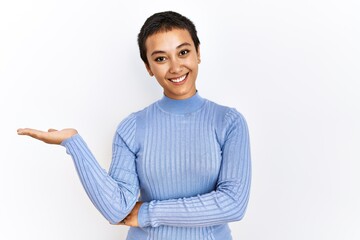 Sticker - Young hispanic woman with short hair standing over isolated background smiling cheerful presenting and pointing with palm of hand looking at the camera.