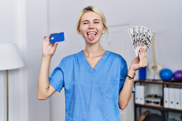 Sticker - Young physiotherapist woman holding credit card and dollars banknotes sticking tongue out happy with funny expression.