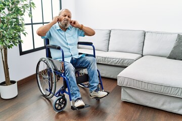 Poster - Handsome senior man sitting on wheelchair at the living room covering ears with fingers with annoyed expression for the noise of loud music. deaf concept.