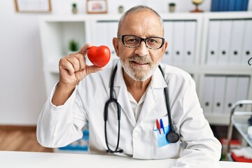 Canvas Print - Mature doctor man holding red heart at the clinic looking positive and happy standing and smiling with a confident smile showing teeth