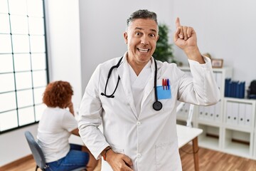 Wall Mural - Middle age doctor man at the clinic with a patient pointing finger up with successful idea. exited and happy. number one.