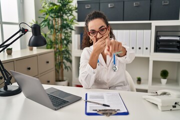 Canvas Print - Young hispanic woman wearing doctor uniform and stethoscope laughing at you, pointing finger to the camera with hand over mouth, shame expression