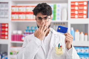 Canvas Print - Hispanic man with beard working at pharmacy drugstore holding credit card covering mouth with hand, shocked and afraid for mistake. surprised expression