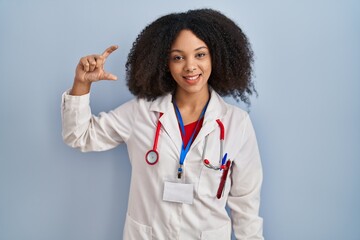 Poster - Young african american woman wearing doctor uniform and stethoscope smiling and confident gesturing with hand doing small size sign with fingers looking and the camera. measure concept.