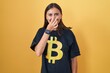 Young hispanic woman wearing bitcoin t shirt smelling something stinky and disgusting, intolerable smell, holding breath with fingers on nose. bad smell