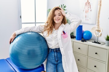 Canvas Print - Young caucasian woman working at rehabilitation clinic holding pilates ball stressed and frustrated with hand on head, surprised and angry face