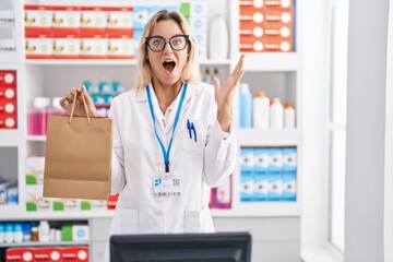 Sticker - Young blonde woman working at pharmacy drugstore holding paper bag celebrating victory with happy smile and winner expression with raised hands