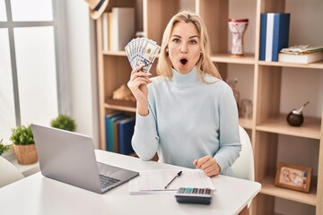 Sticker - Young caucasian woman using laptop holding dollars banknotes scared and amazed with open mouth for surprise, disbelief face