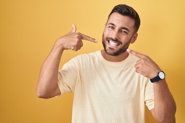 Canvas Print - Handsome hispanic man standing over yellow background smiling cheerful showing and pointing with fingers teeth and mouth. dental health concept.