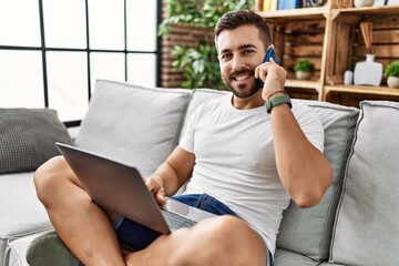 Poster - Young hispanic man using laptop and talking on the smartphone at home