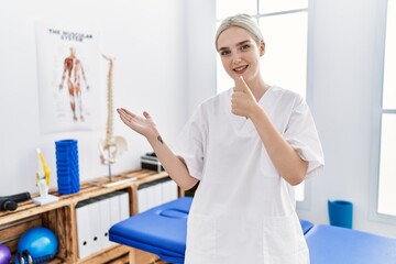 Poster - Young caucasian woman working at pain recovery clinic showing palm hand and doing ok gesture with thumbs up, smiling happy and cheerful