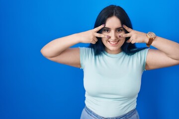 Canvas Print - Young modern girl with blue hair standing over blue background doing peace symbol with fingers over face, smiling cheerful showing victory