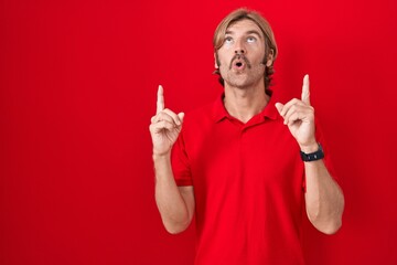 Poster - Caucasian man with mustache standing over red background amazed and surprised looking up and pointing with fingers and raised arms.