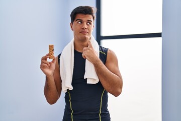 Canvas Print - Hispanic man eating protein bar as healthy energy snack serious face thinking about question with hand on chin, thoughtful about confusing idea