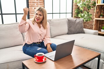 Poster - Young hispanic woman using laptop sitting on the sofa at home angry and mad raising fist frustrated and furious while shouting with anger. rage and aggressive concept.