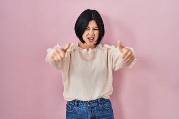 Sticker - Young asian woman standing over pink background approving doing positive gesture with hand, thumbs up smiling and happy for success. winner gesture.