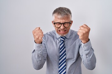 Sticker - Hispanic business man with grey hair wearing glasses angry and mad raising fists frustrated and furious while shouting with anger. rage and aggressive concept.