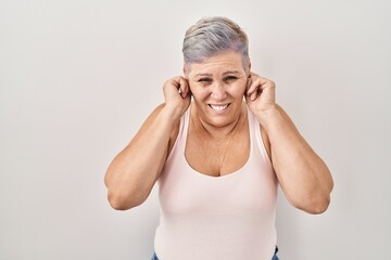 Sticker - Middle age caucasian woman standing over white background covering ears with fingers with annoyed expression for the noise of loud music. deaf concept.