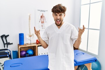 Canvas Print - Young arab man working at pain recovery clinic shouting with crazy expression doing rock symbol with hands up. music star. heavy concept.