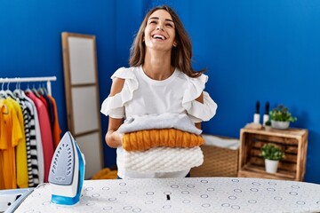 Poster - Young hispanic woman ironing clothes at laundry room holding folded sweaters smiling and laughing hard out loud because funny crazy joke.