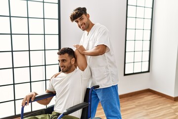 Canvas Print - Two hispanic men physiotherapist and patient sitting on wheelchair having rehab session stretching arm at clinic