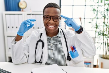 Poster - Young african doctor man holding syringe at the hospital smiling pointing to head with one finger, great idea or thought, good memory