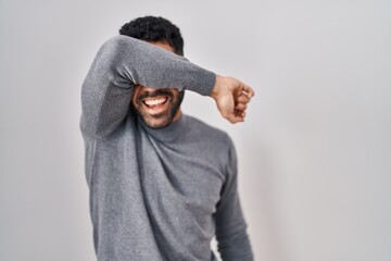 Wall Mural - Hispanic man with beard standing over white background covering eyes with arm smiling cheerful and funny. blind concept.