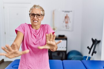 Poster - Middle age blonde woman at pain recovery clinic afraid and terrified with fear expression stop gesture with hands, shouting in shock. panic concept.