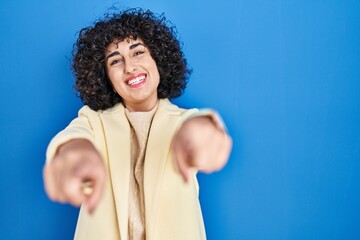 Wall Mural - Young brunette woman with curly hair standing over blue background pointing to you and the camera with fingers, smiling positive and cheerful