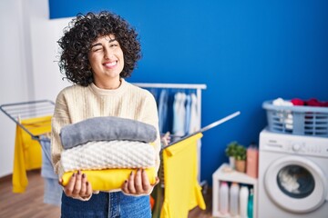 Poster - Young brunette woman with curly hair holding clean laundry winking looking at the camera with sexy expression, cheerful and happy face.