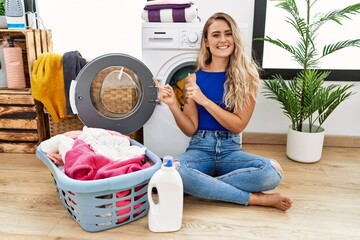 Canvas Print - Young beautiful woman doing laundry sitting by wicker basket smiling and looking at the camera pointing with two hands and fingers to the side.