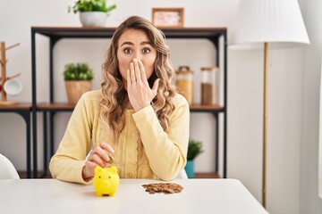 Poster - Young caucasian woman sitting on the table with piggy bank and coins covering mouth with hand, shocked and afraid for mistake. surprised expression