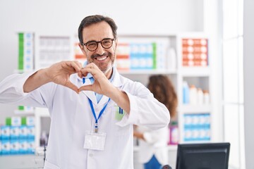 Wall Mural - Middle age hispanic man working at pharmacy drugstore smiling in love doing heart symbol shape with hands. romantic concept.