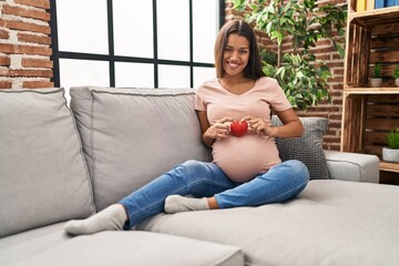 Sticker - Young latin woman pregnant holding heart sitting on sofa at home