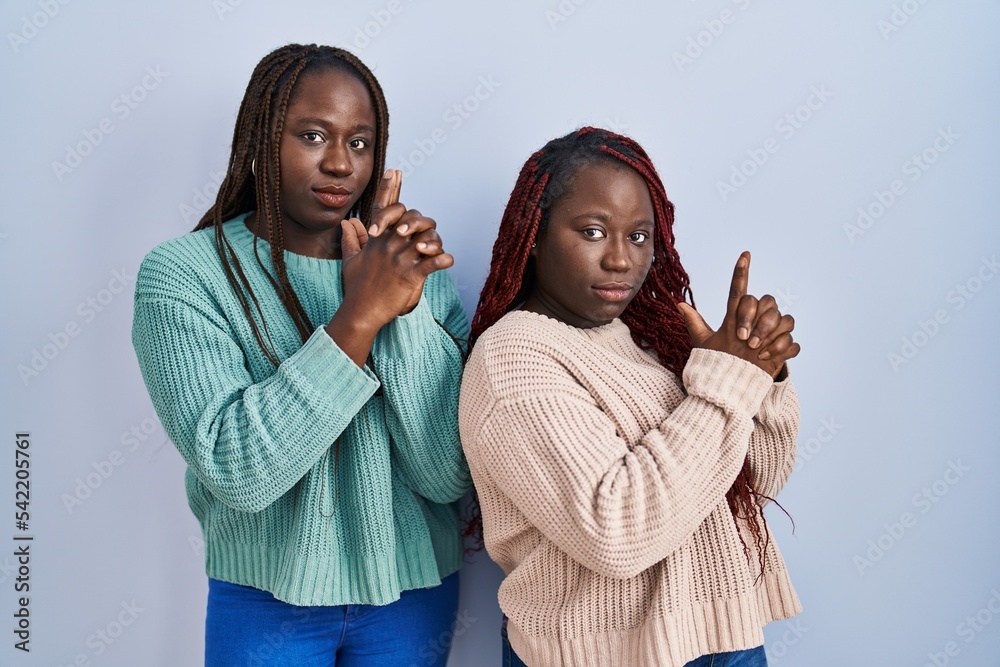 Two african woman standing over blue background holding symbolic gun with hand gesture, playing killing shooting weapons, angry face