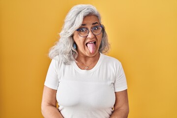 Poster - Middle age woman with grey hair standing over yellow background sticking tongue out happy with funny expression. emotion concept.