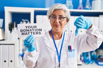 Wall Mural - Middle age woman with grey hair working at scientist laboratory holding your donation matters banner with angry face, negative sign showing dislike with thumbs down, rejection concept