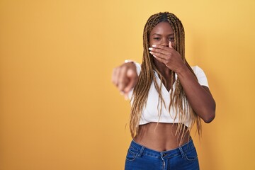 Wall Mural - African american woman with braided hair standing over yellow background laughing at you, pointing finger to the camera with hand over mouth, shame expression