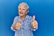 © Krakenimages.com - Senior woman with grey hair standing over blue background approving doing positive gesture with hand, thumbs up smiling and happy for success. winner gesture.